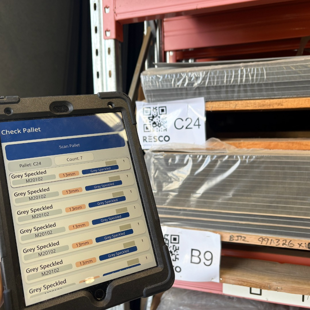 The foreground of this image shows an IPAD and custom made software used to track stock in a warehouse. In the background pallets with QR codes are Shown